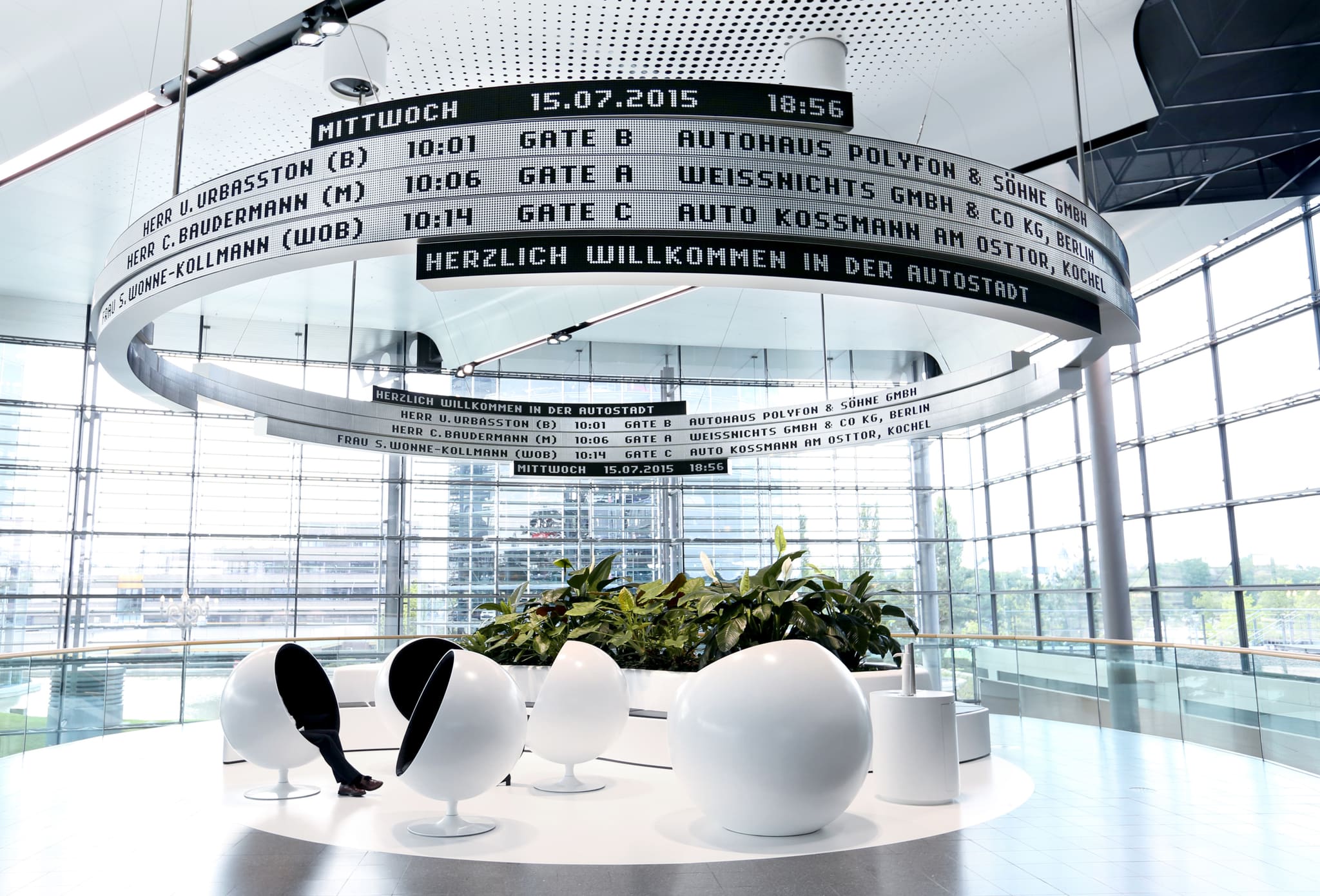 Ring-shaped display board below the hall ceiling, with white ball chairs, a planted bed and a glass façade