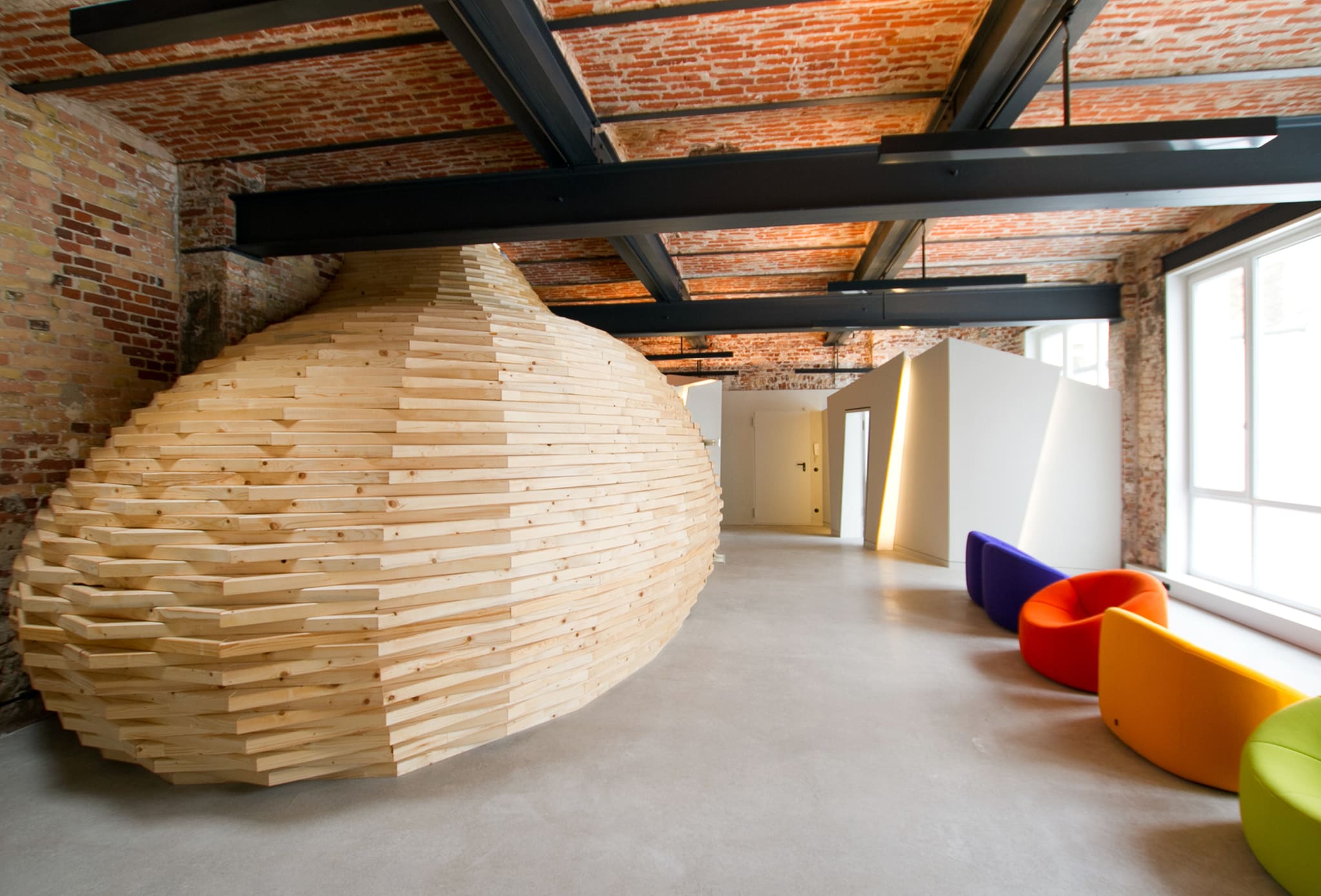 Stacked timber volume in a loft, brick vaulted ceiling with black steel beams, concrete floor, colourful round seats