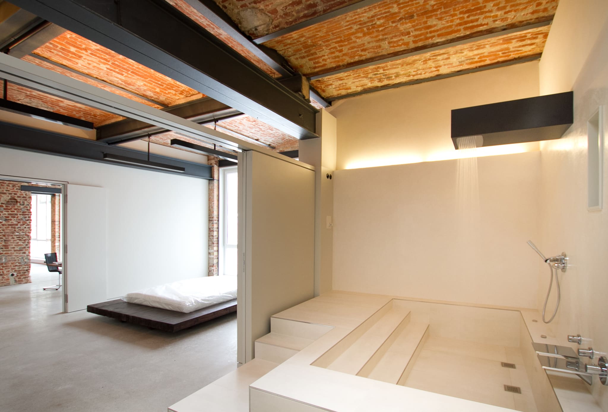 Bed on dark timber platform behind white sliding wall, at right sunken tub with steps and shower box