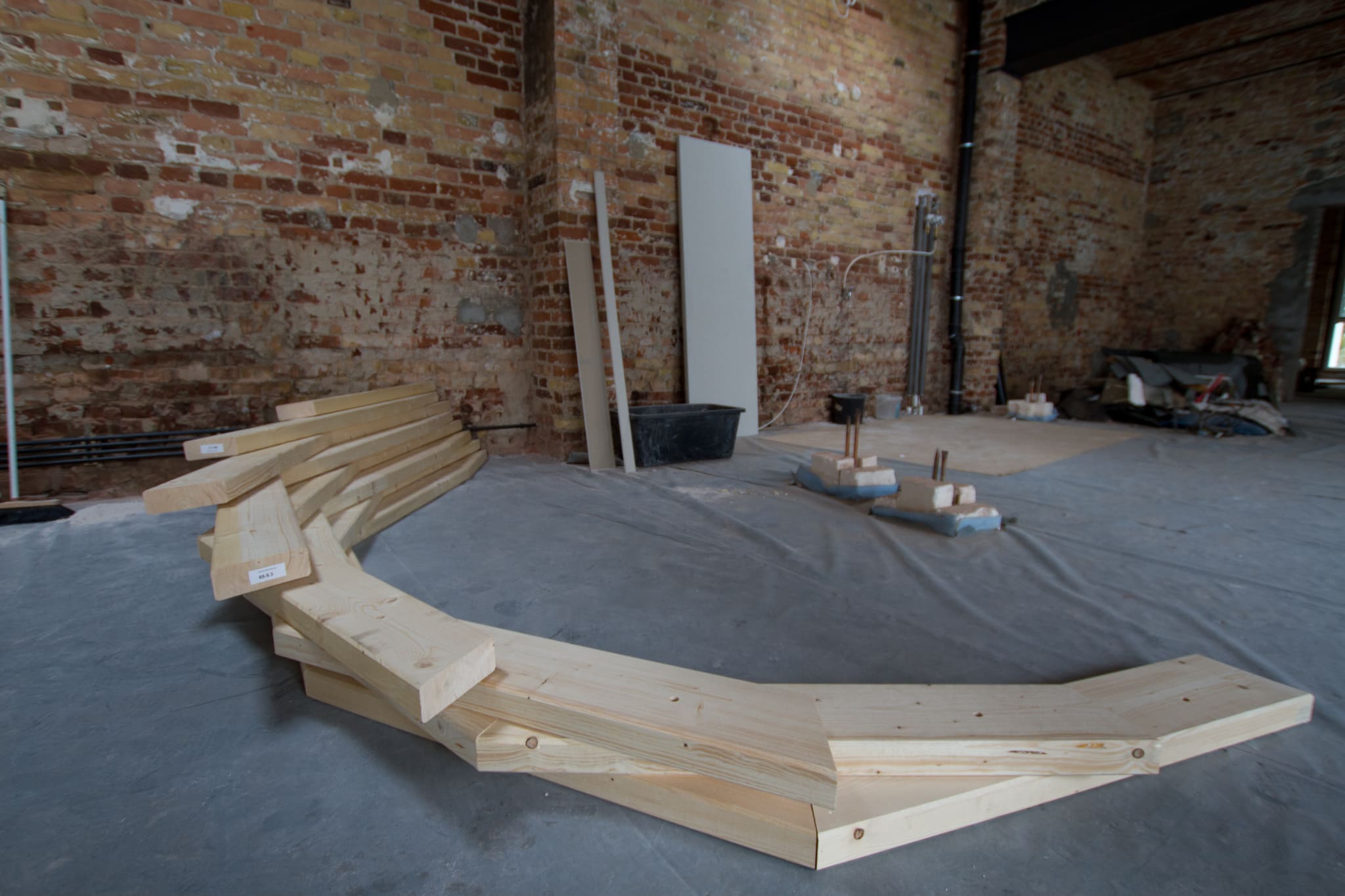 First layer of the timber boards for the cocoon, laid as a curved arc on the site floor in front of a brick wall