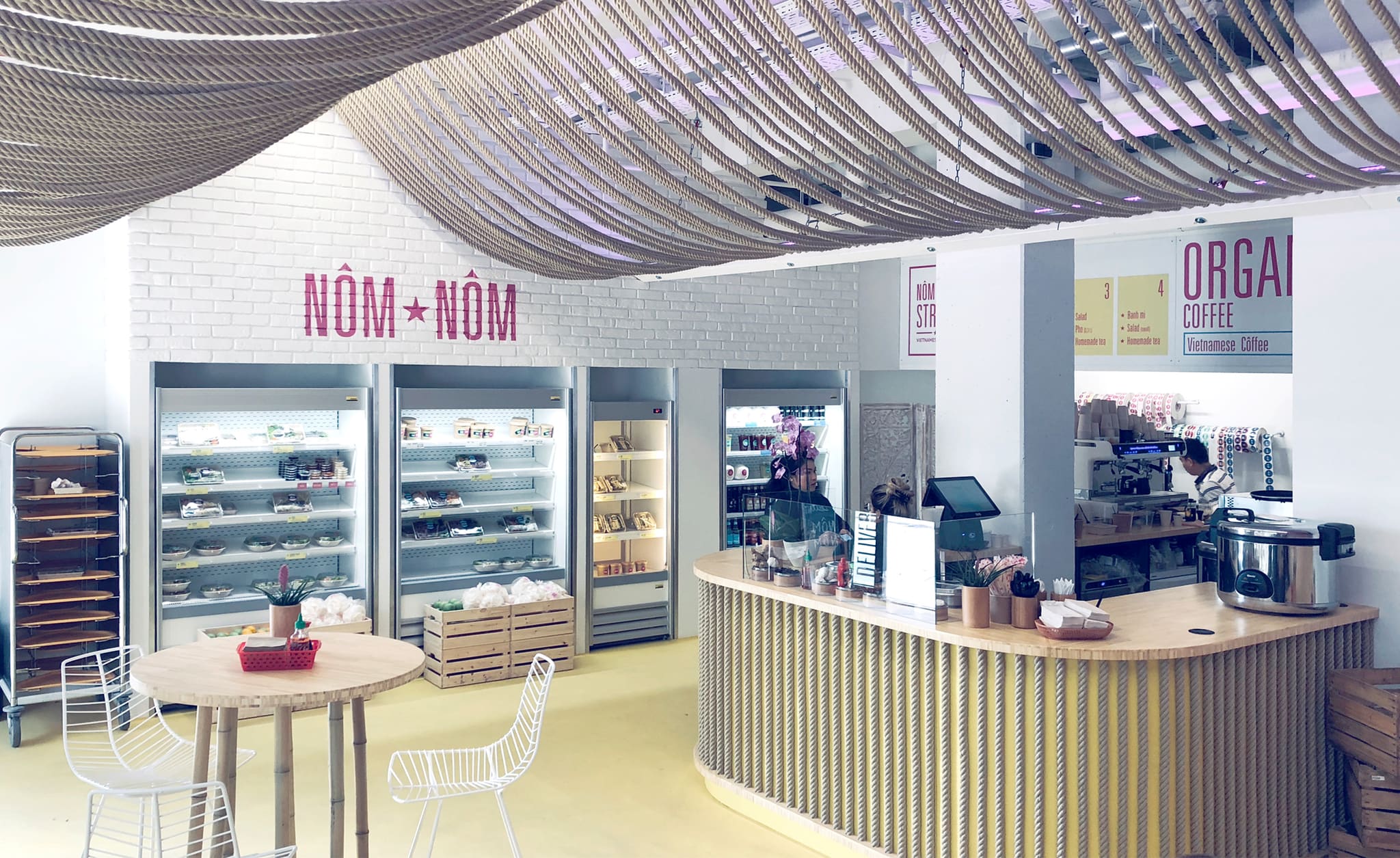 Dining area with a natural rope ceiling, a white brick wall with Nôm Nôm lettering, chilled display cabinets and a curved counter