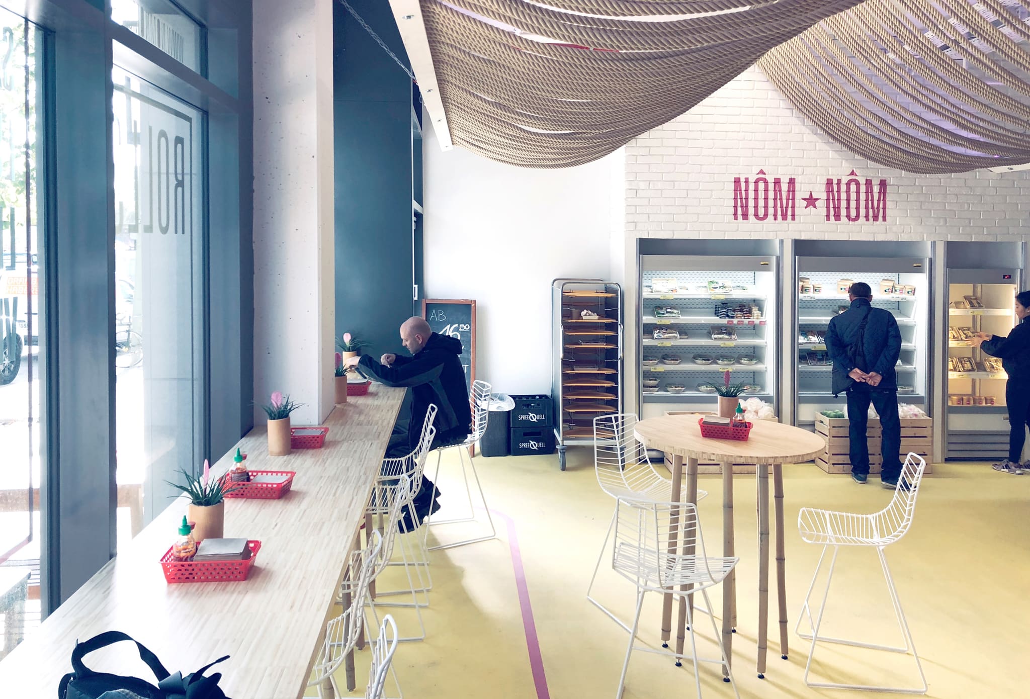 Long bamboo counter at the shop window with white wire chairs, chilled display units and a white brick wall behind
