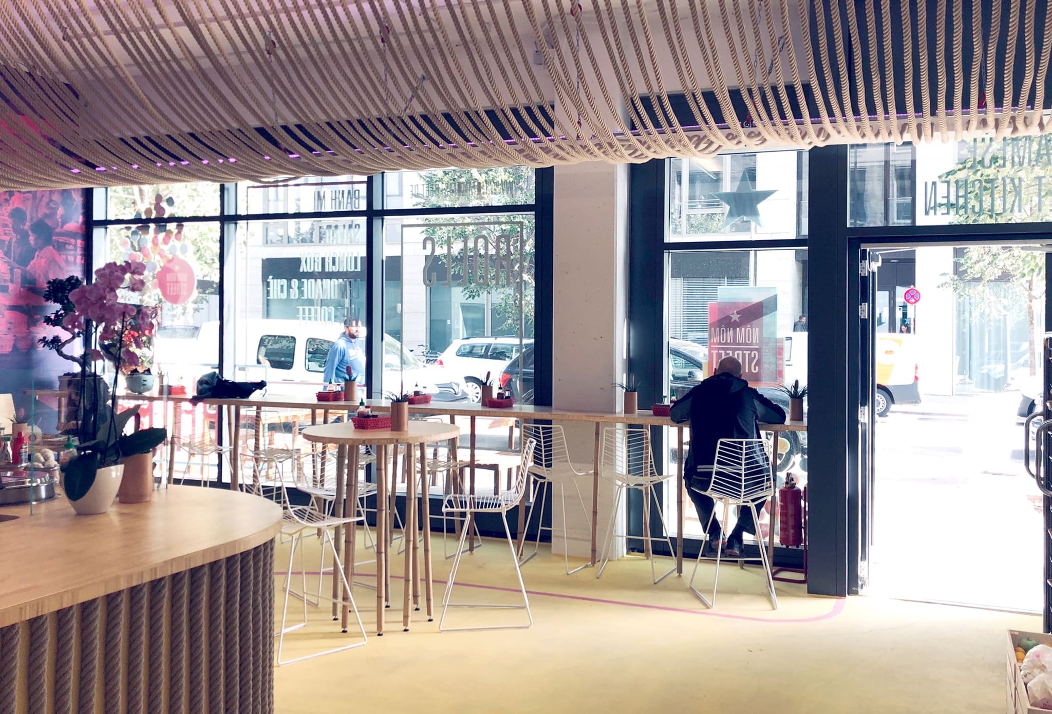 View to the shopfront: curved rope ceiling, bamboo high tables with white wire chairs, open entrance door, the street beyond