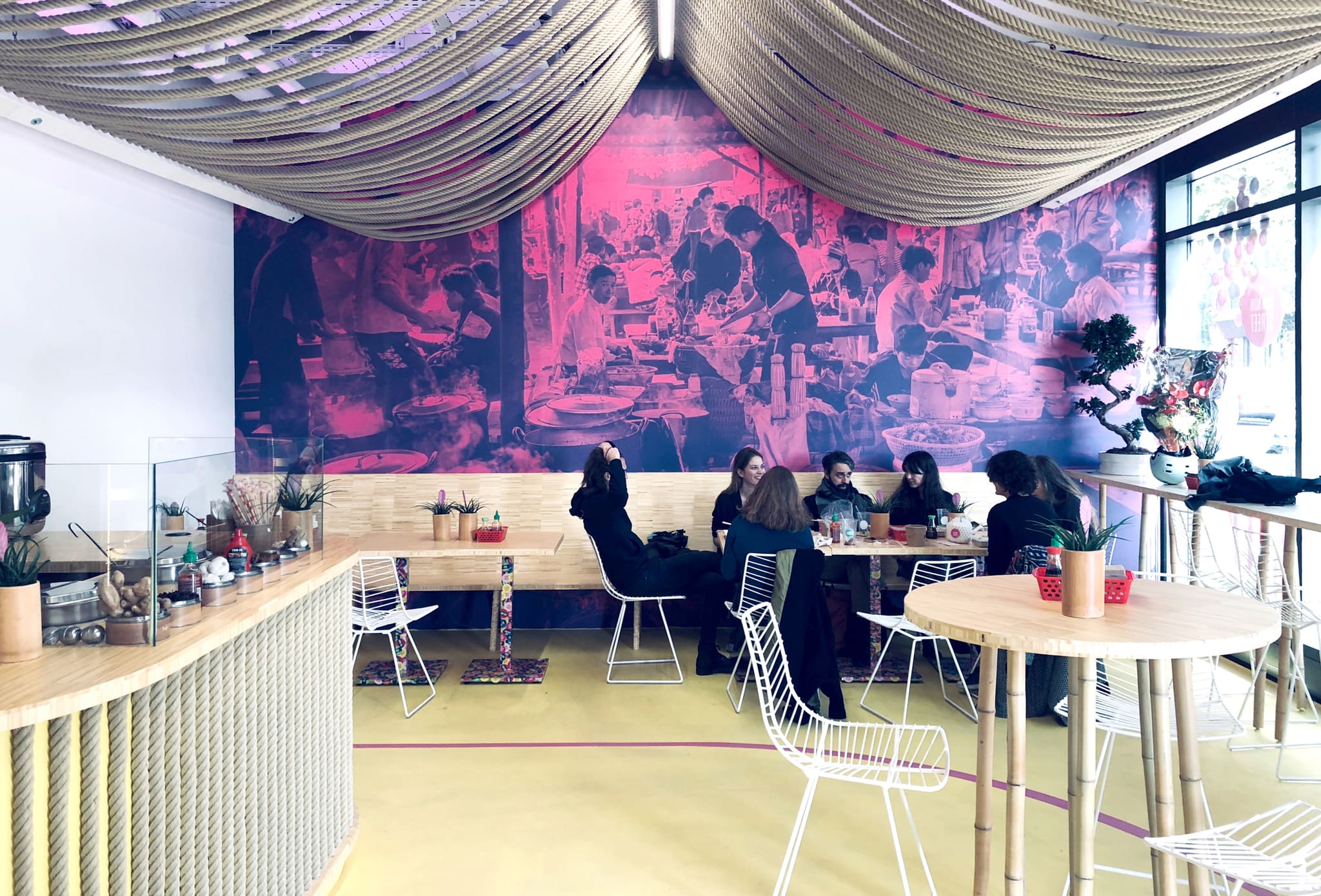 Seating area with a magenta-tinted mural of a market kitchen, bamboo tables, white wire chairs, low-hanging rope ceiling, several guests