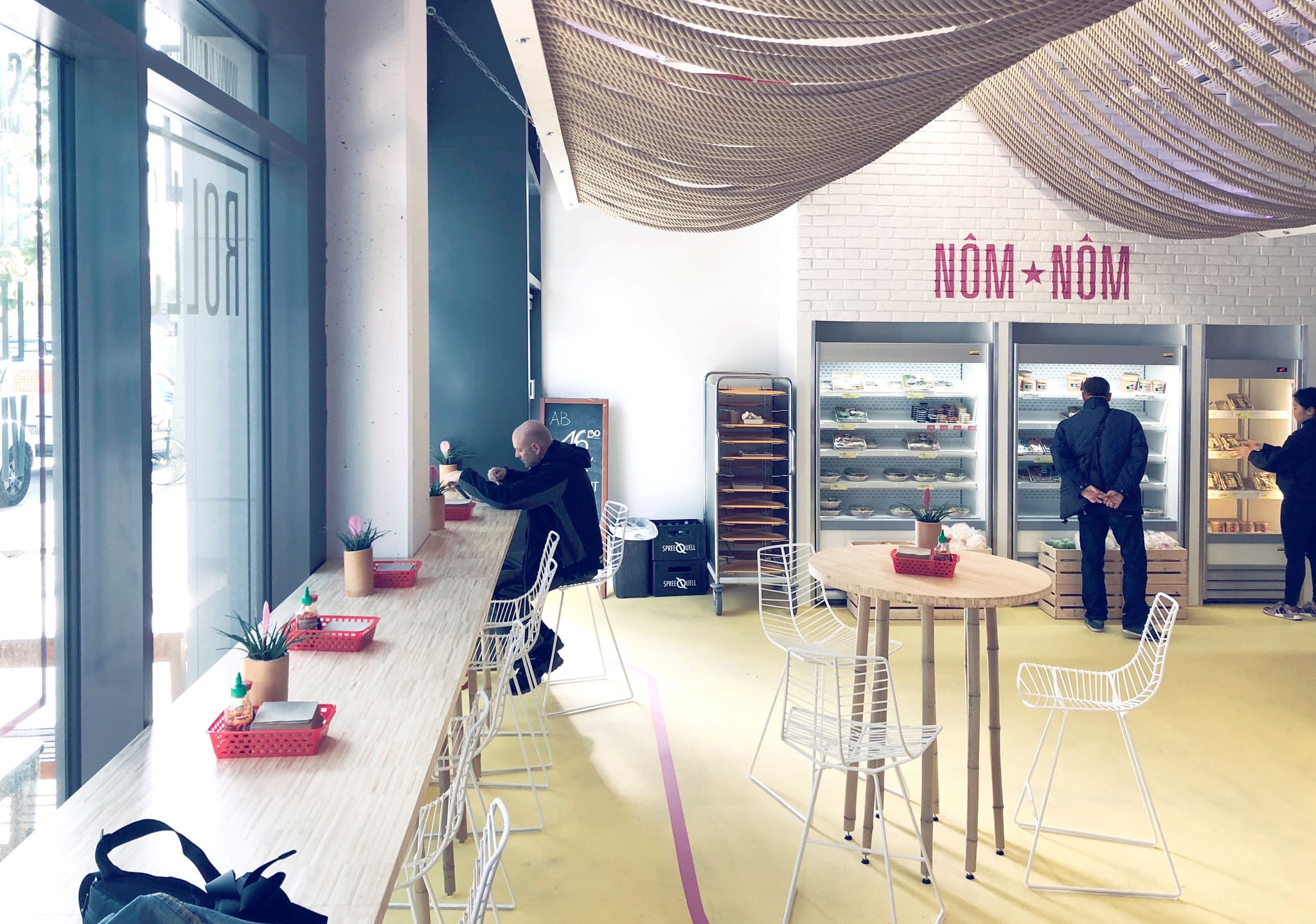 Long window bar with bar stools, behind it chilled display cabinets and the Nôm Nôm lettering below the natural rope ceiling
