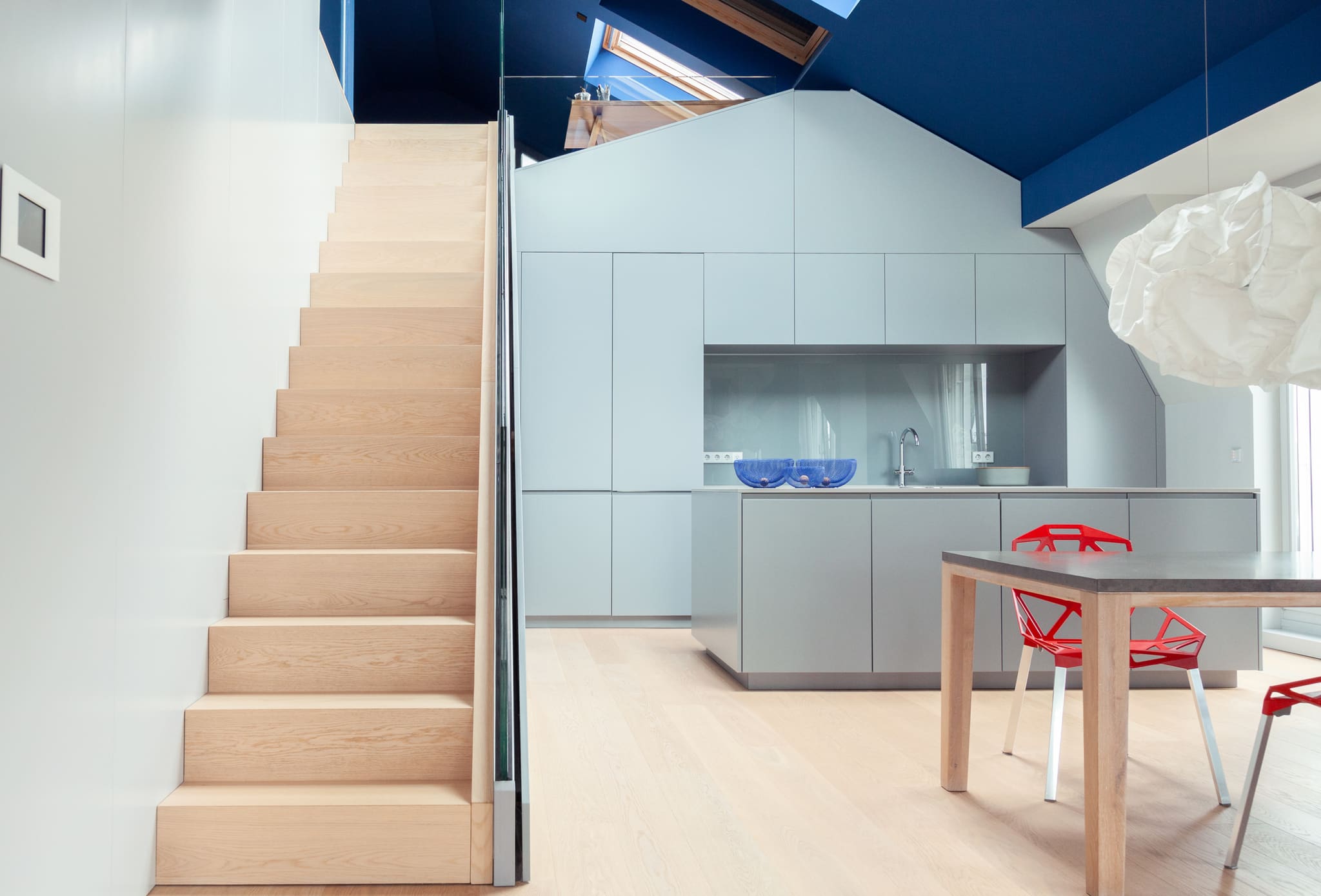 Open living space, pale oak stair with glass balustrade, handle-less light grey kitchen behind, dark blue roof space with skylights
