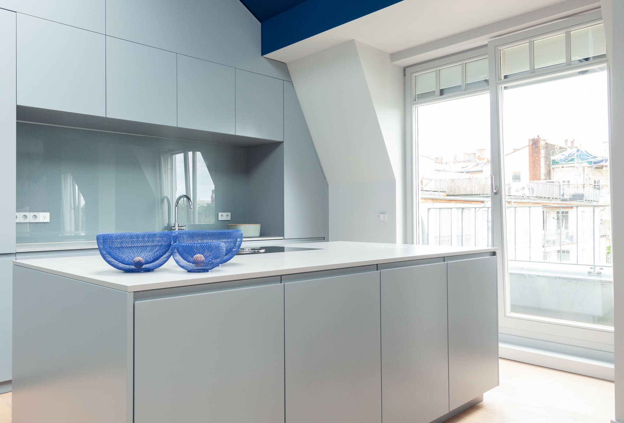 Light grey kitchen with glass splashback and island, two blue wire bowls on the worktop, tall window onto neighbouring roofs