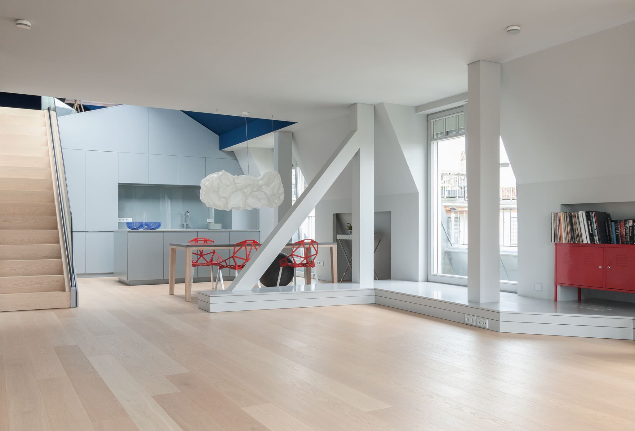 Wide living room with pale oak boards, angled white columns and struts, kitchen and dining table with red chairs behind