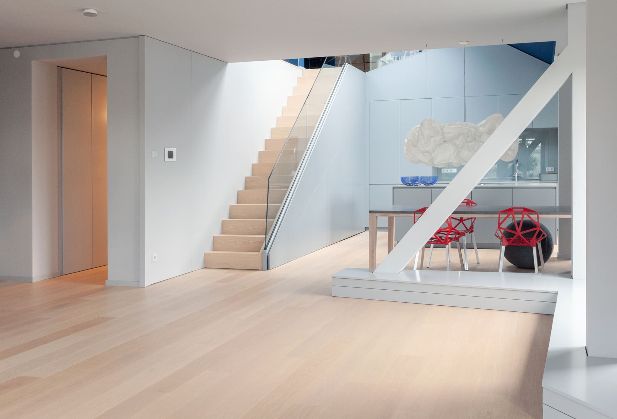 View across oak boards to the stair with glass balustrade, white built-in wall with flush door, dining table at right