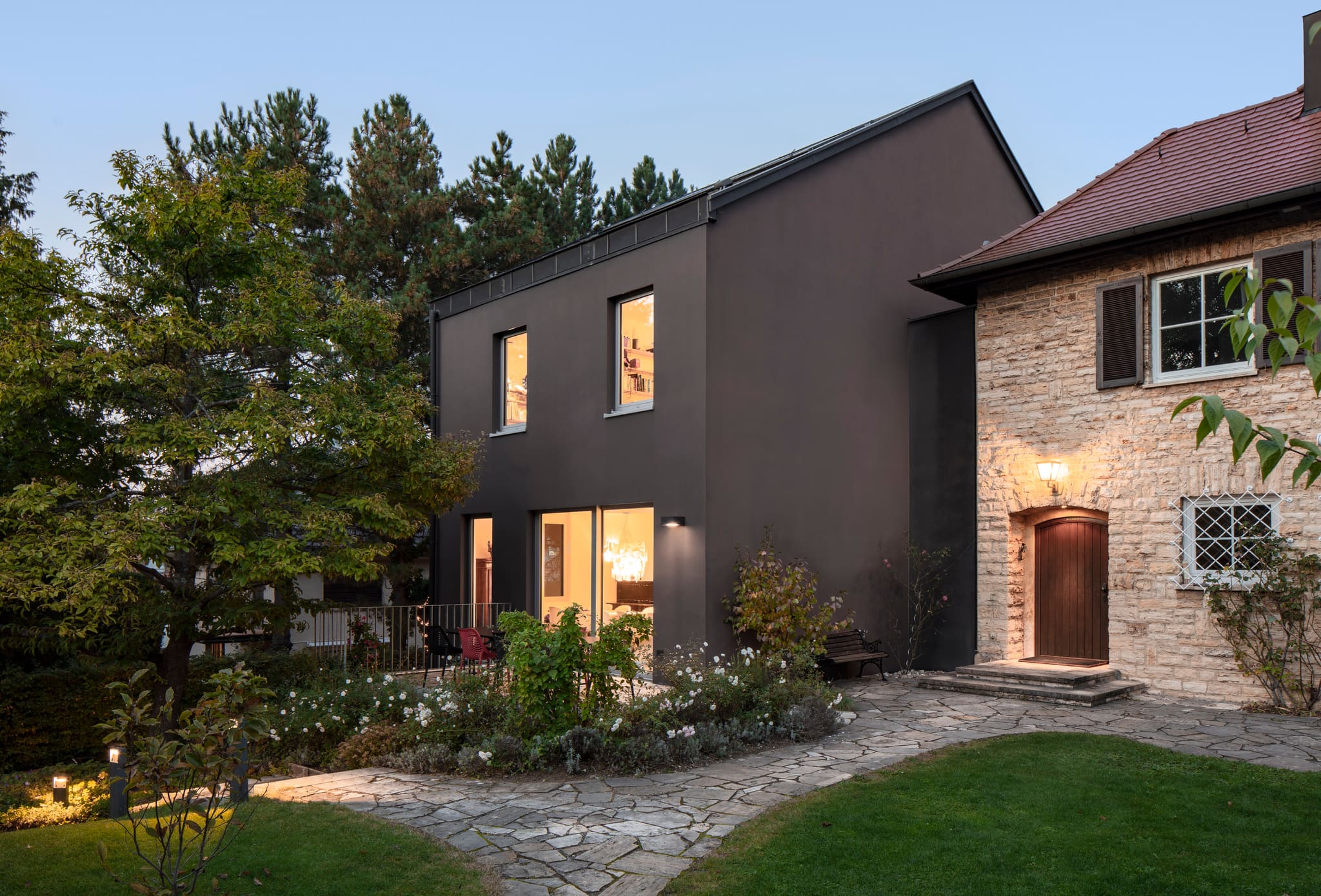 Dark rendered new house at dusk with lit windows, the old stone house at right, rose beds and flagstone path