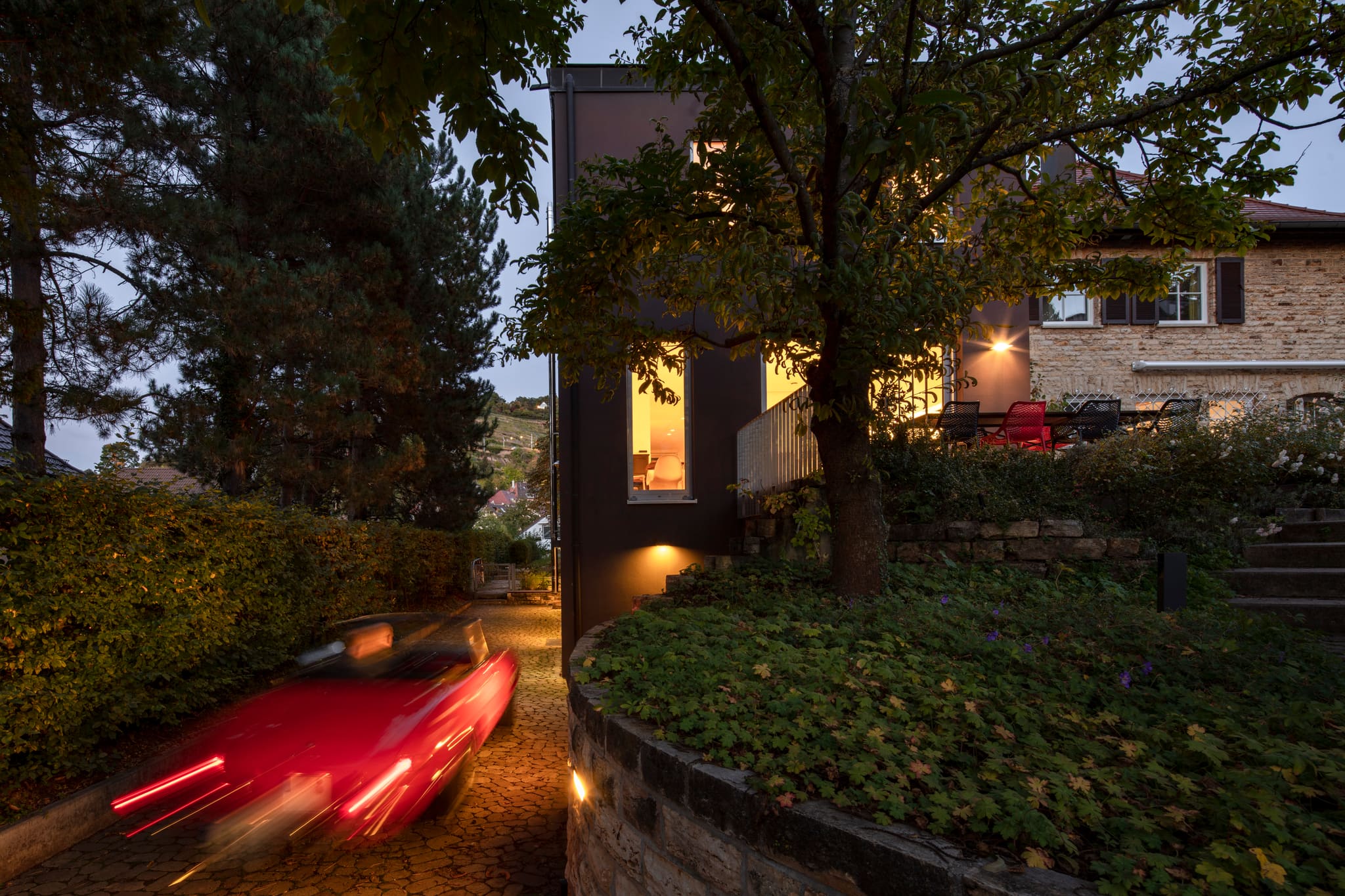 Nachtaufnahme der schmalen Zufahrt zwischen dunklem Neubau und Natursteinhaus, beleuchtete Fenster, Trockenmauer, wegfahrendes rotes Auto
