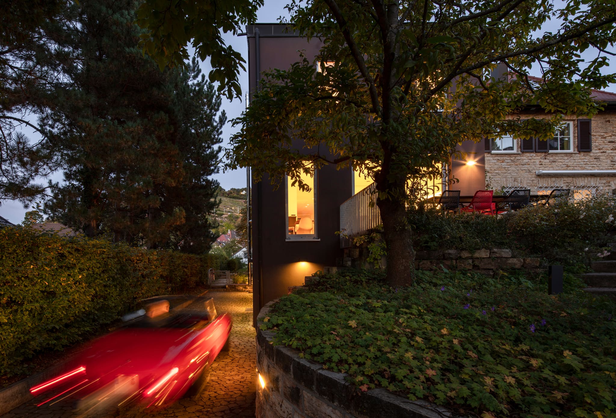 Night view of the narrow drive between dark new build and stone house, lit windows, dry stone wall, red car