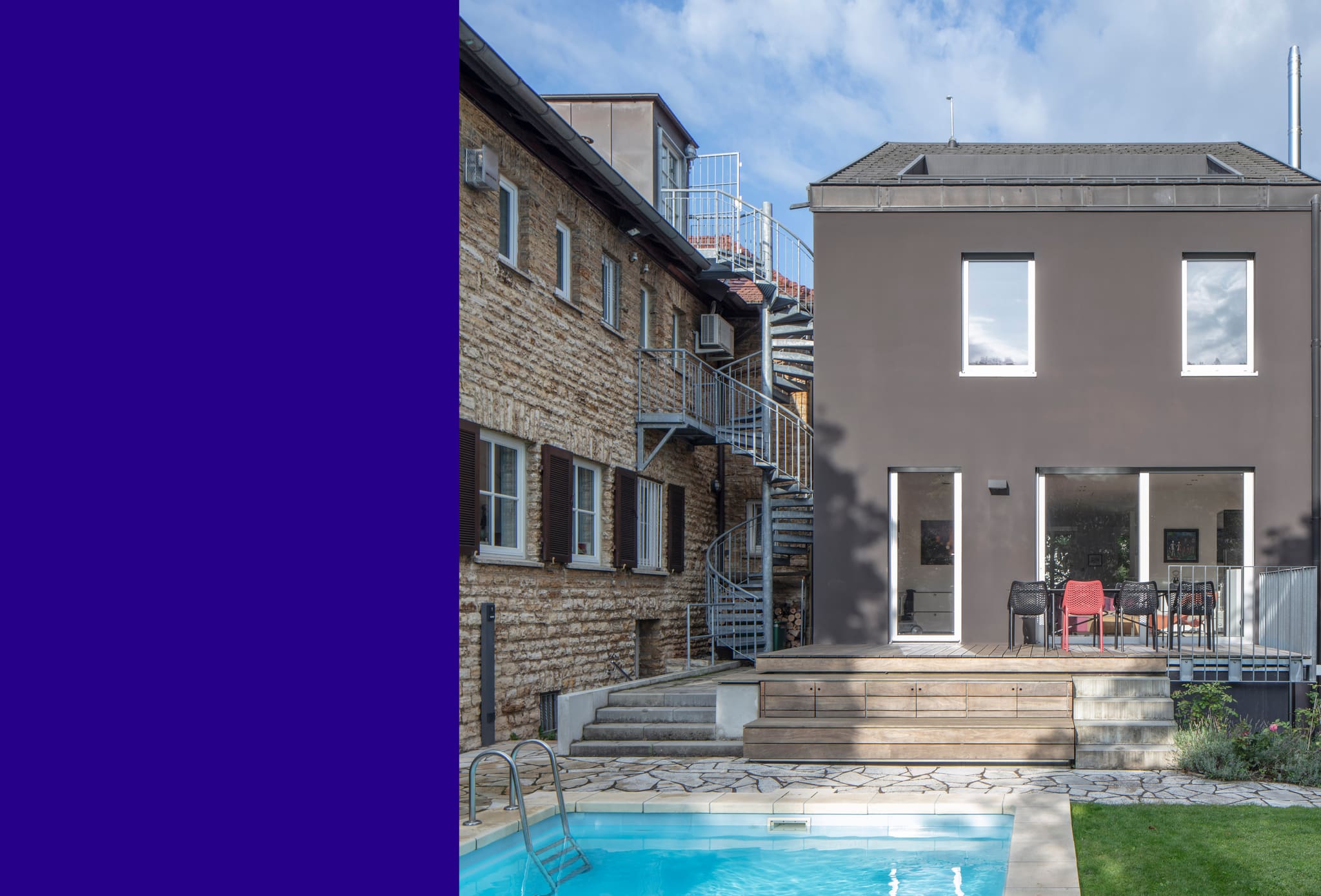 Garden side by day: stone old building with spiral staircase, rendered new build with timber deck, pool in the foreground
