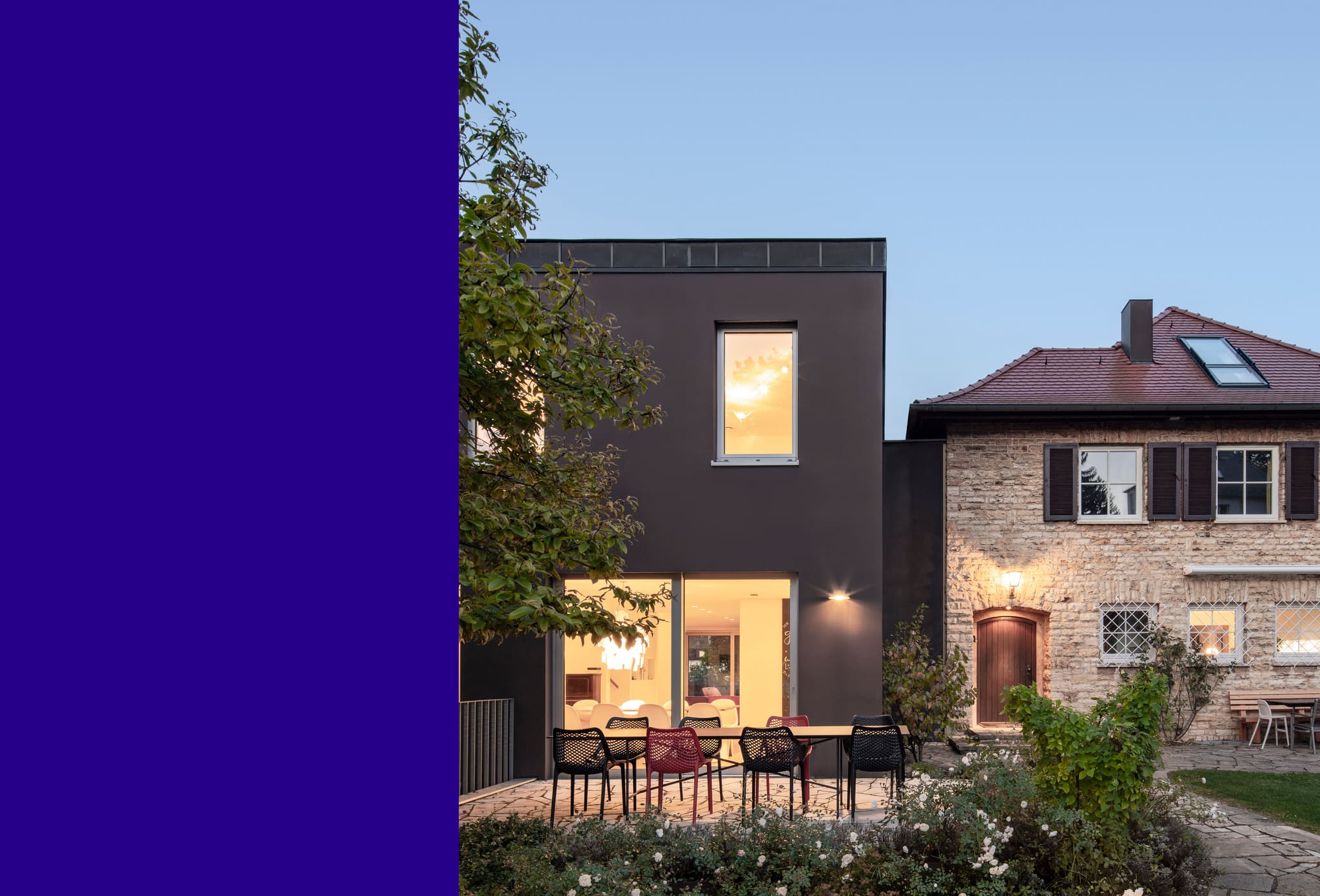 Garden façade at dusk, lit living and dining area behind sliding glazing, outdoor table with black and red chairs