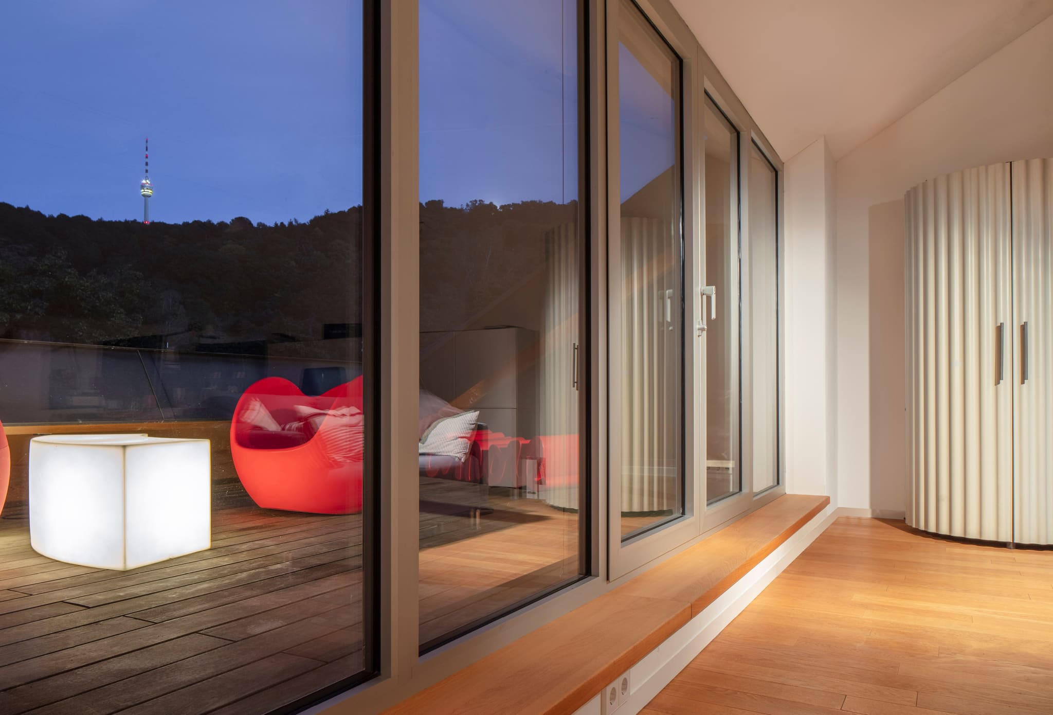 Room with timber floor and window seat, terrace with red armchair and glowing cube stool, wooded slope with television tower