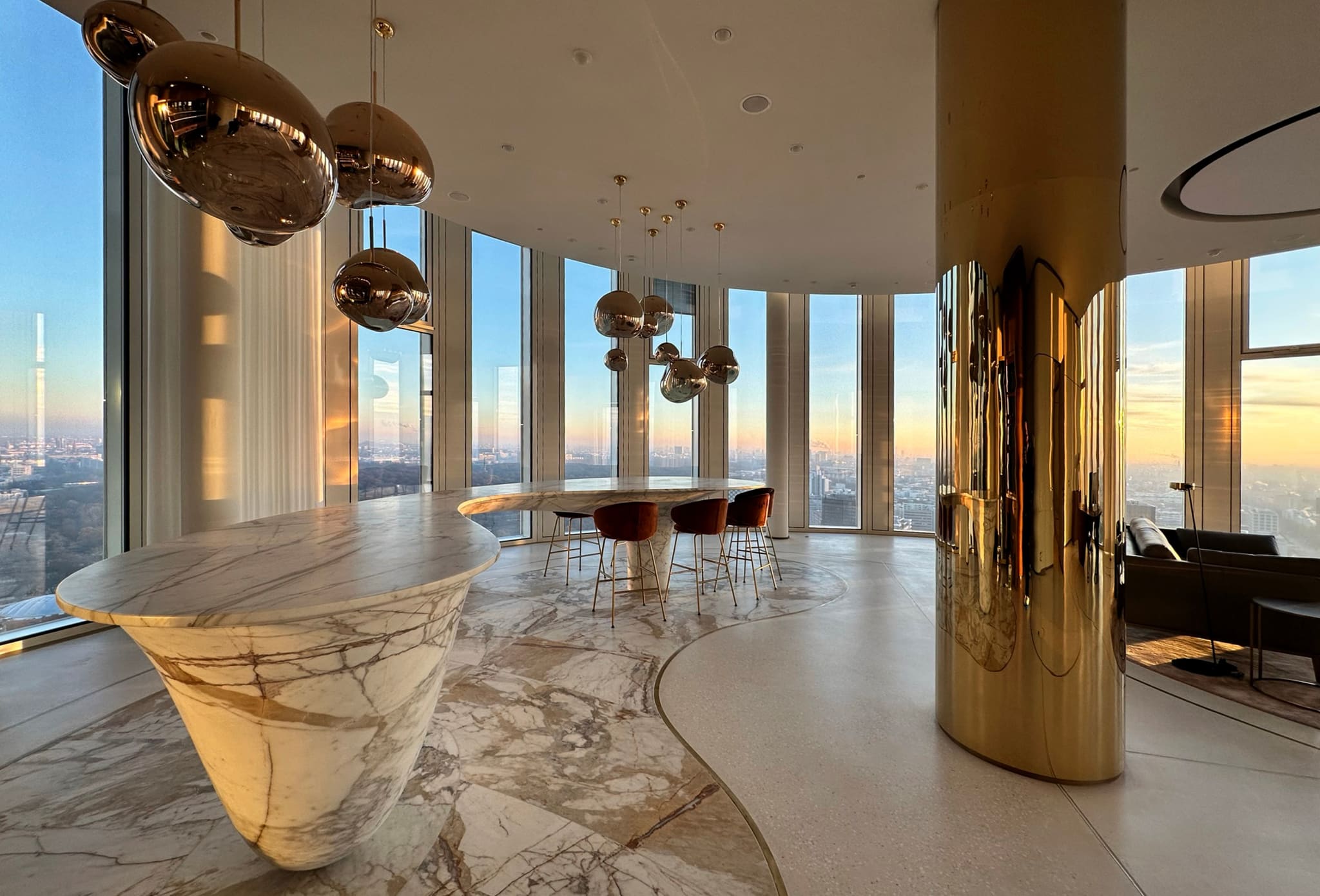 Penthouse room at sunset, curved marble counter and round marble table with velvet stools, brass-clad column, panoramic windows