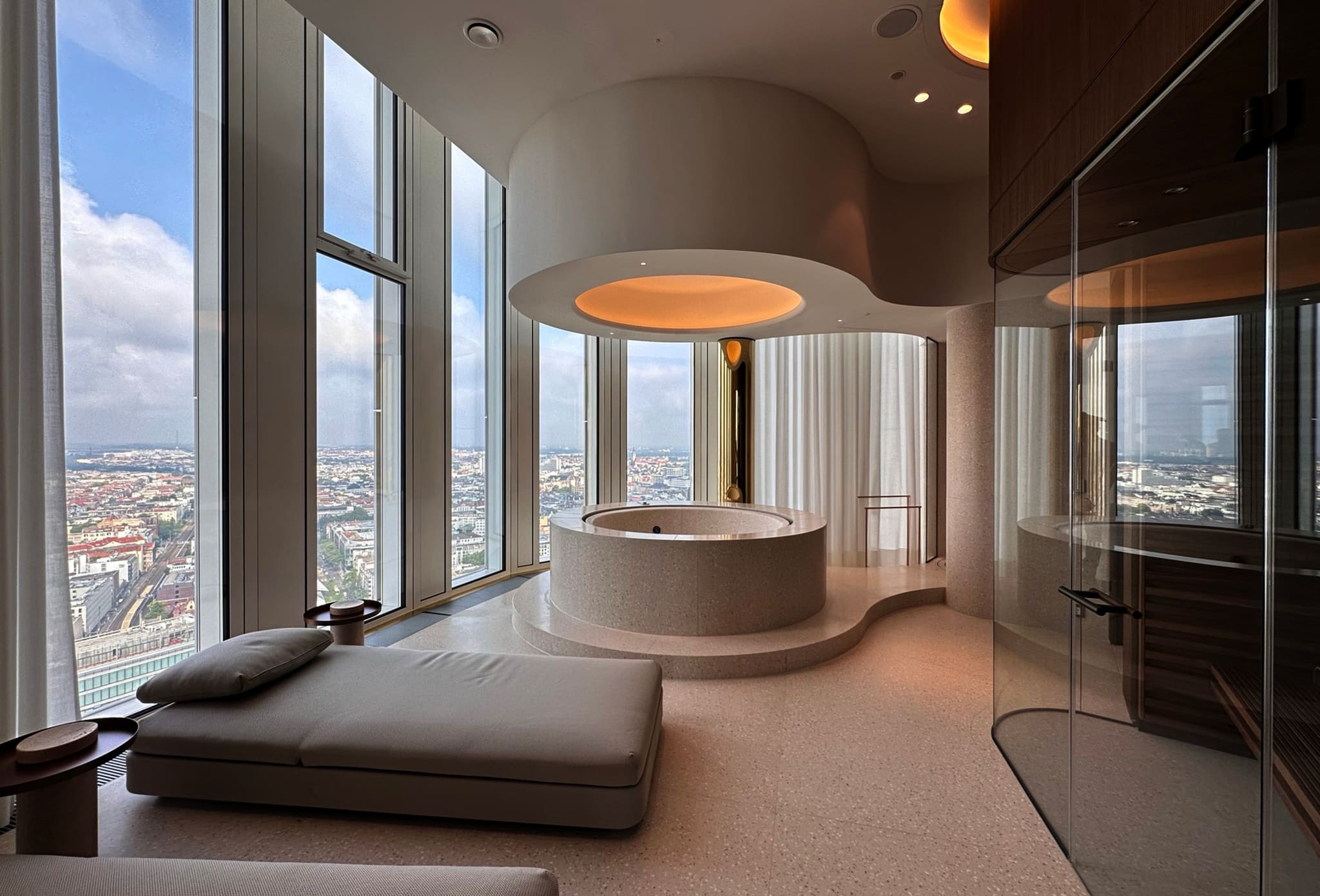 Spa with a round terrazzo tub on a plinth, backlit ceiling opening, daybed at the window, sauna behind glass at right