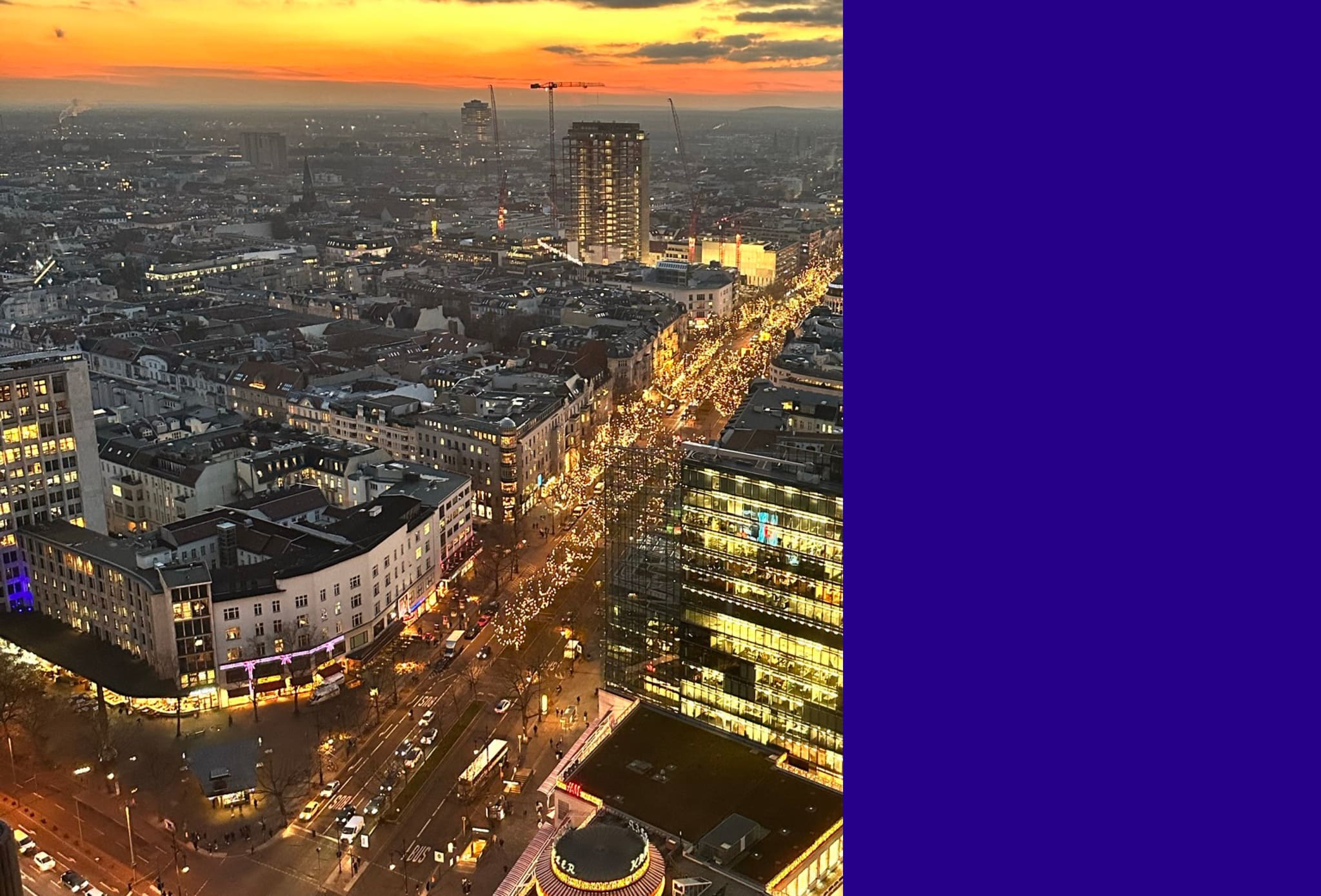 Aerial view at dusk, illuminated boulevard with strings of lights, old rooftops, glass-fronted office building, construction crane on the horizon