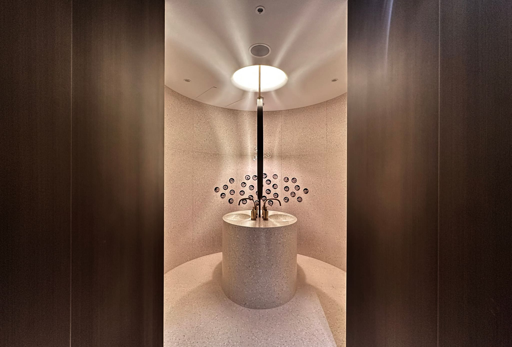 Round terrazzo washstand with two basins and brass taps, wall niches arranged in a semicircle behind, dark timber panelling framing the view