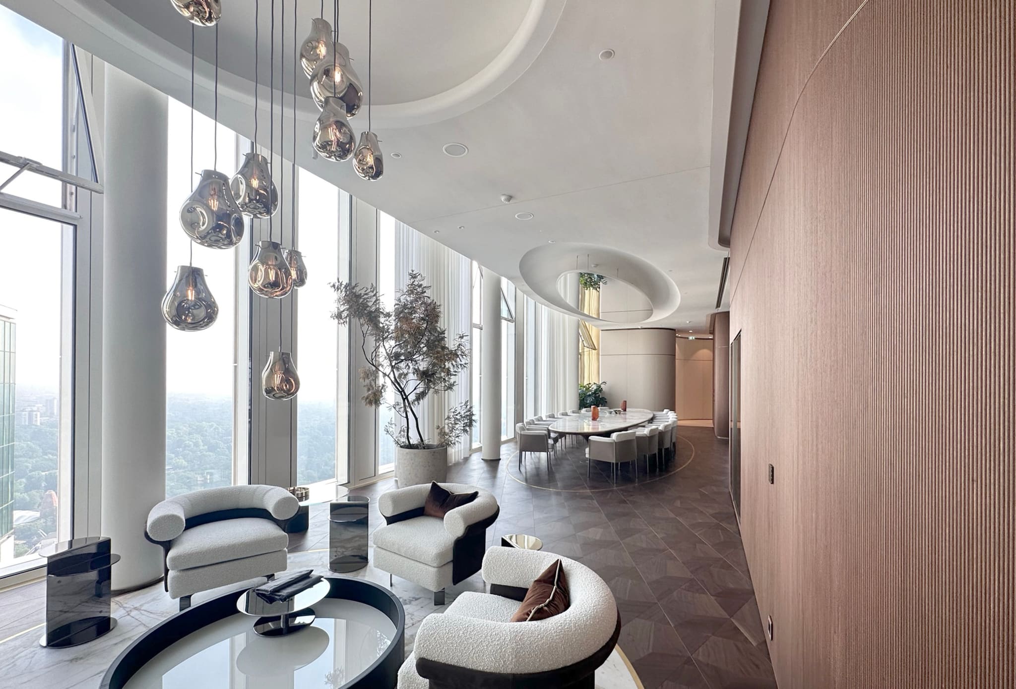 Lounge with white bouclé armchairs and glass tables, cluster of mirrored glass pendants, ribbed timber wall at right, long dining table behind
