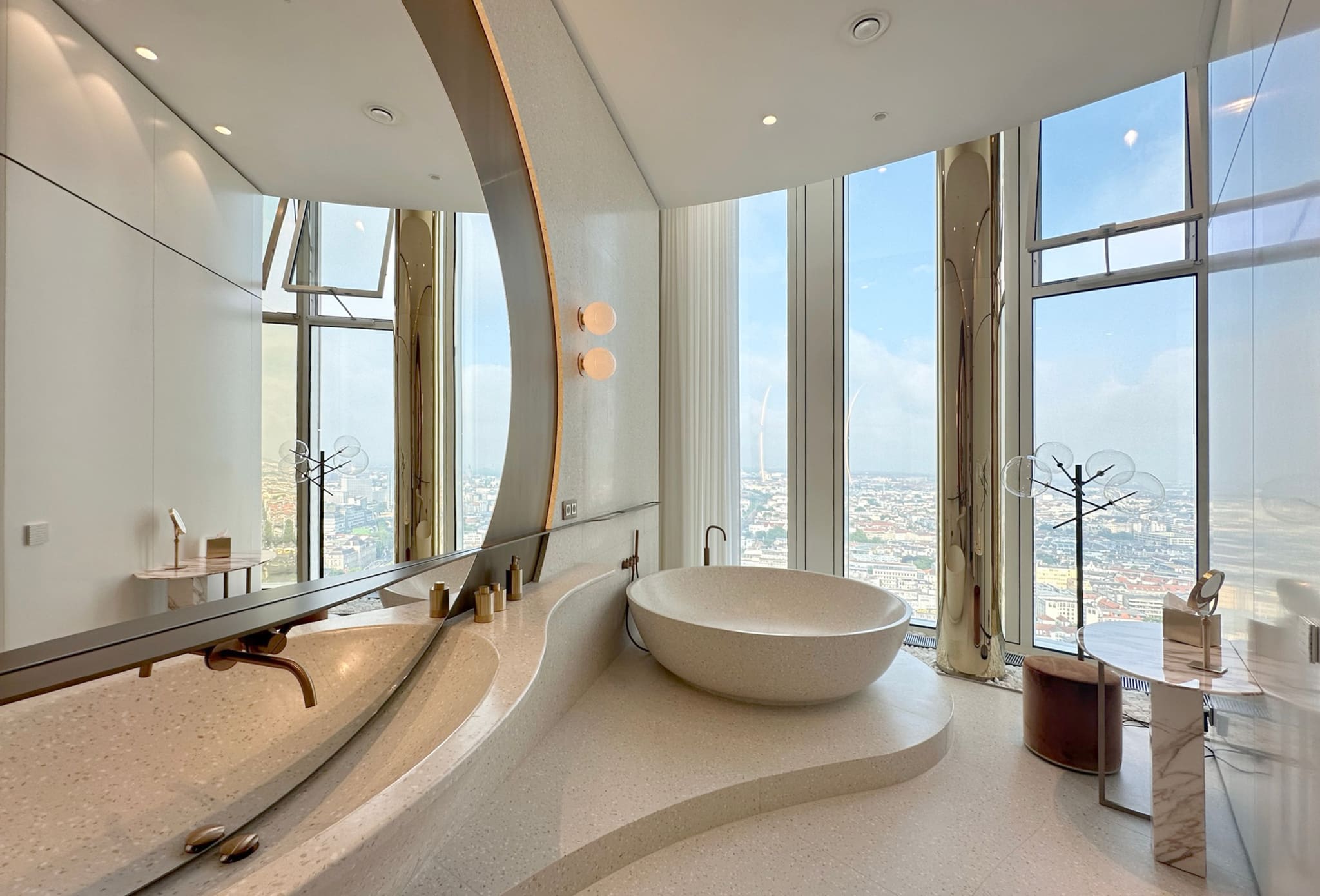 Bathroom with an oval tub on a terrazzo plinth at the window, long washstand with bronze taps, large curved mirror