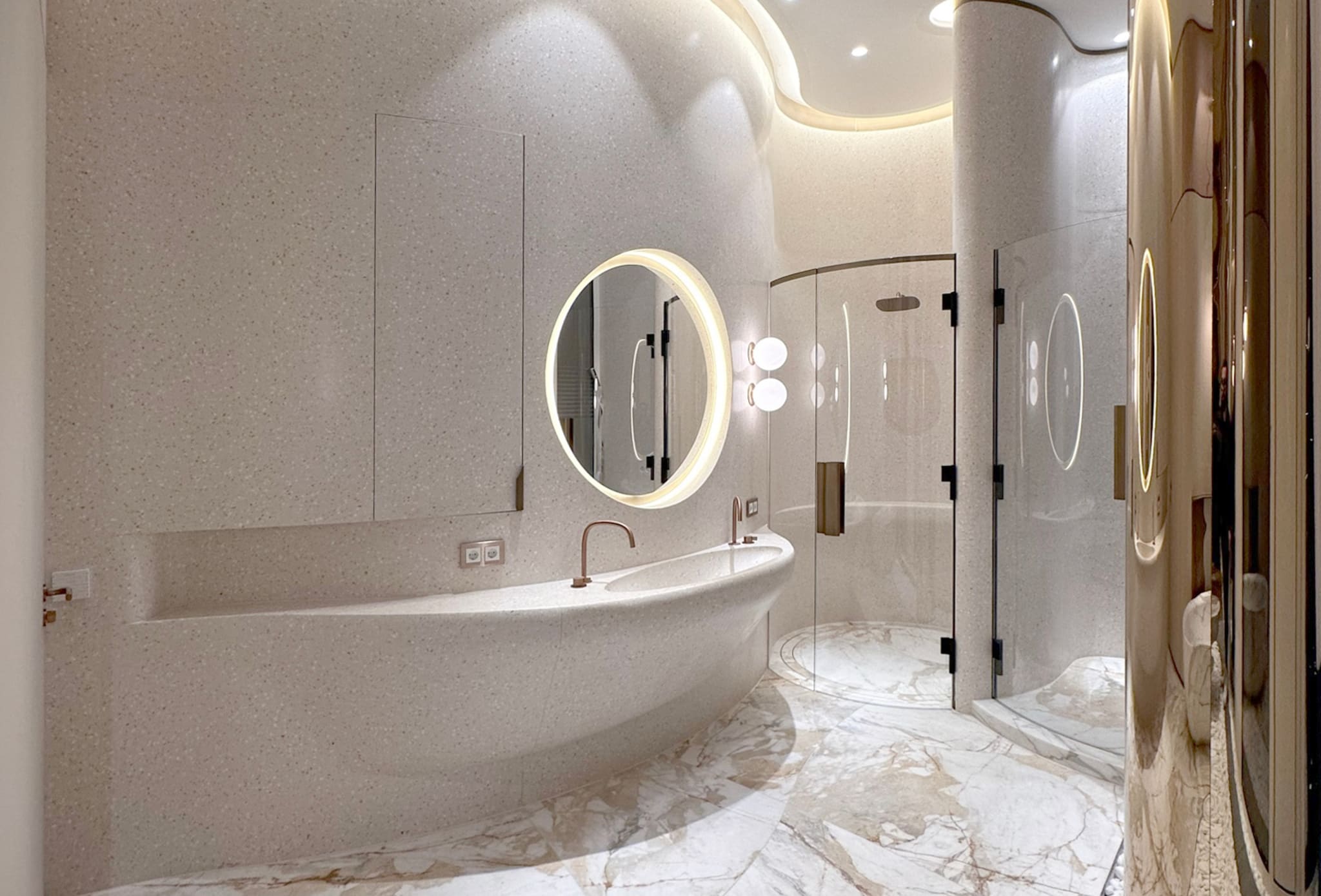 Guest bathroom with monolithic terrazzo washstand, lit round mirror, curved glass shower and marble floor with warm veining