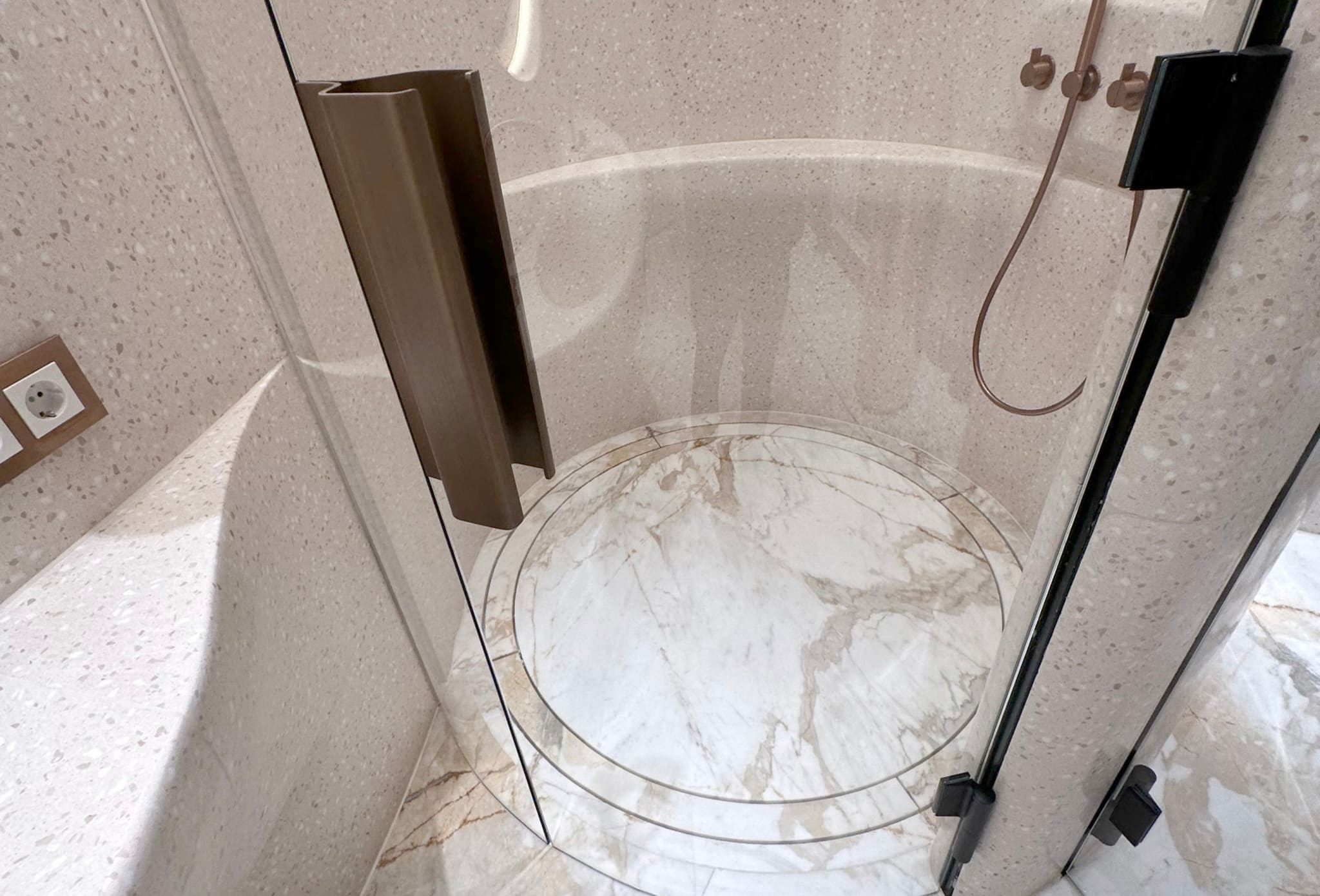 Close-up of a round shower enclosure with curved glass door, bronze handle, marble floor with a circular joint and terrazzo walls