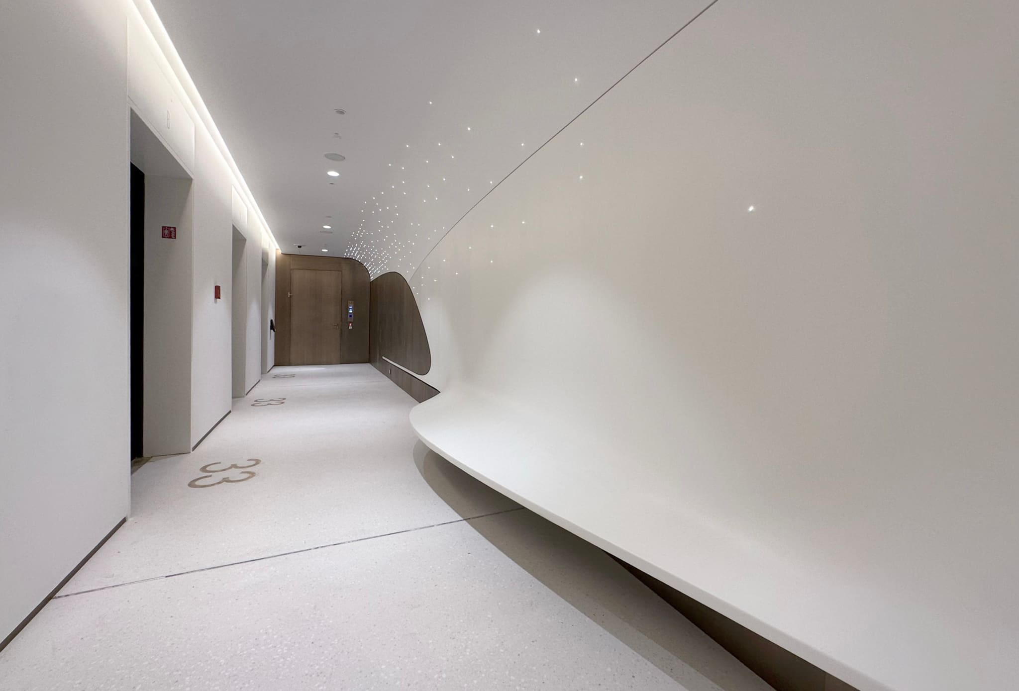 White corridor with a curved wall shell, hundreds of small ceiling light points, terrazzo floor and timber-clad doors at the end