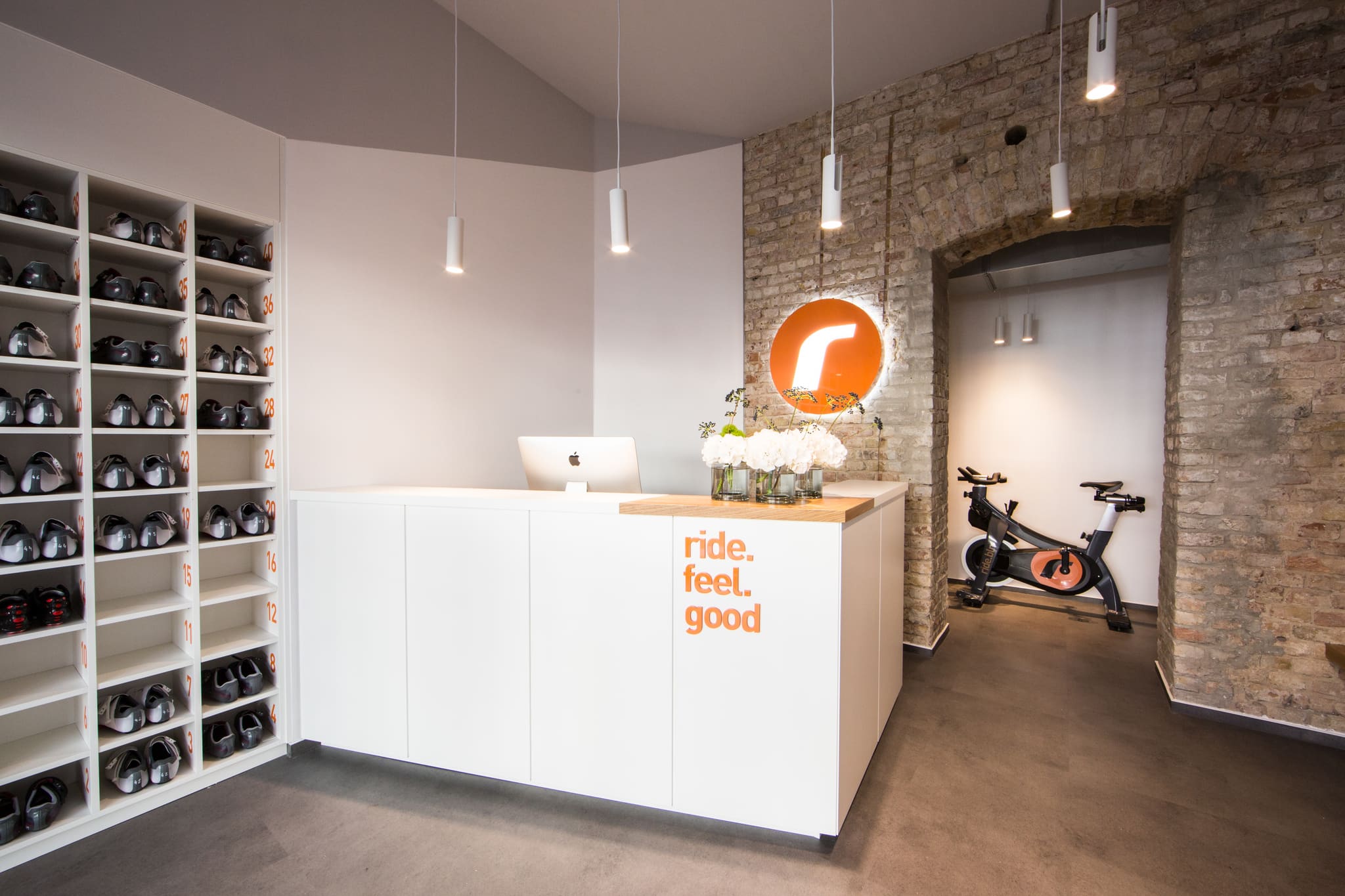 White reception counter with a wooden top in front of an exposed brick wall, an illuminated round logo panel behind it, shelving with cycling shoes on the left