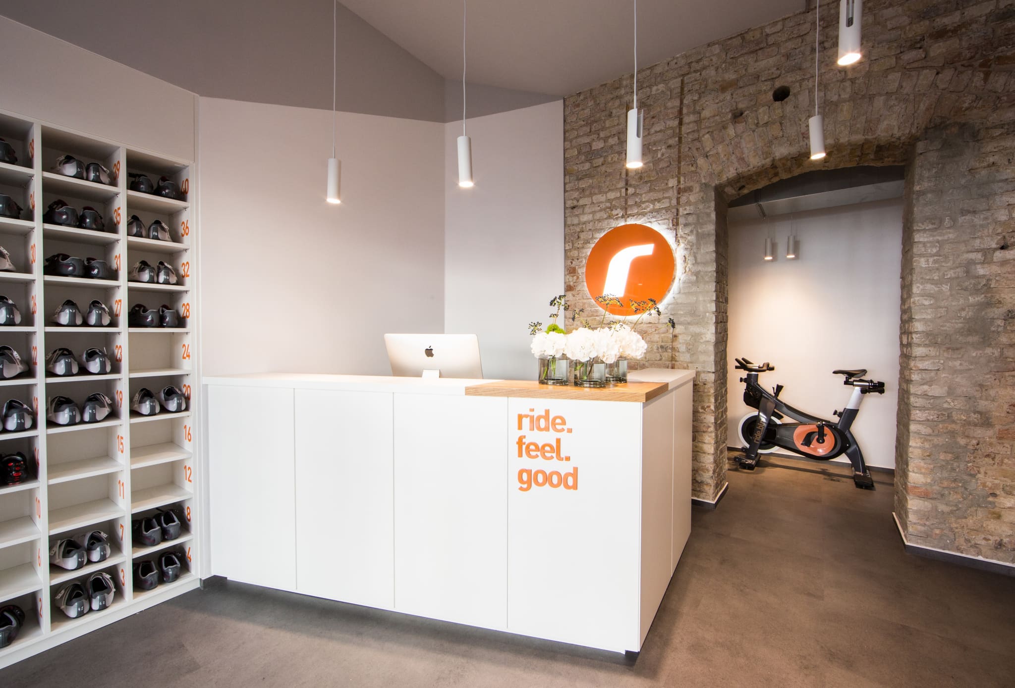 White reception counter with oak top, shelf wall of cycling shoes, old brick wall with arch and an indoor bike