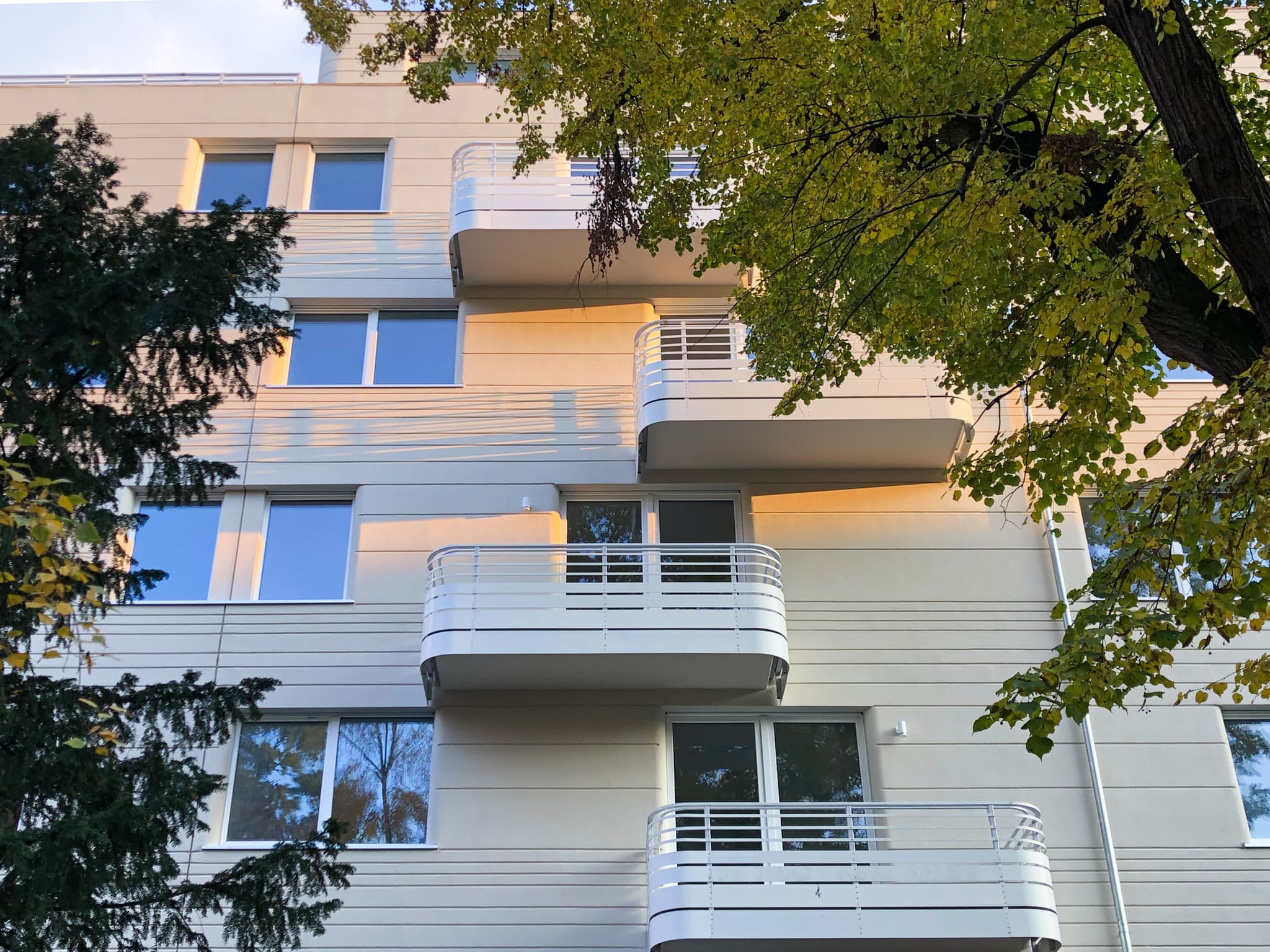 Blick von unten auf die helle Fassade mit gerundeten, integrierten Balkonen, gerahmt von Baumzweigen