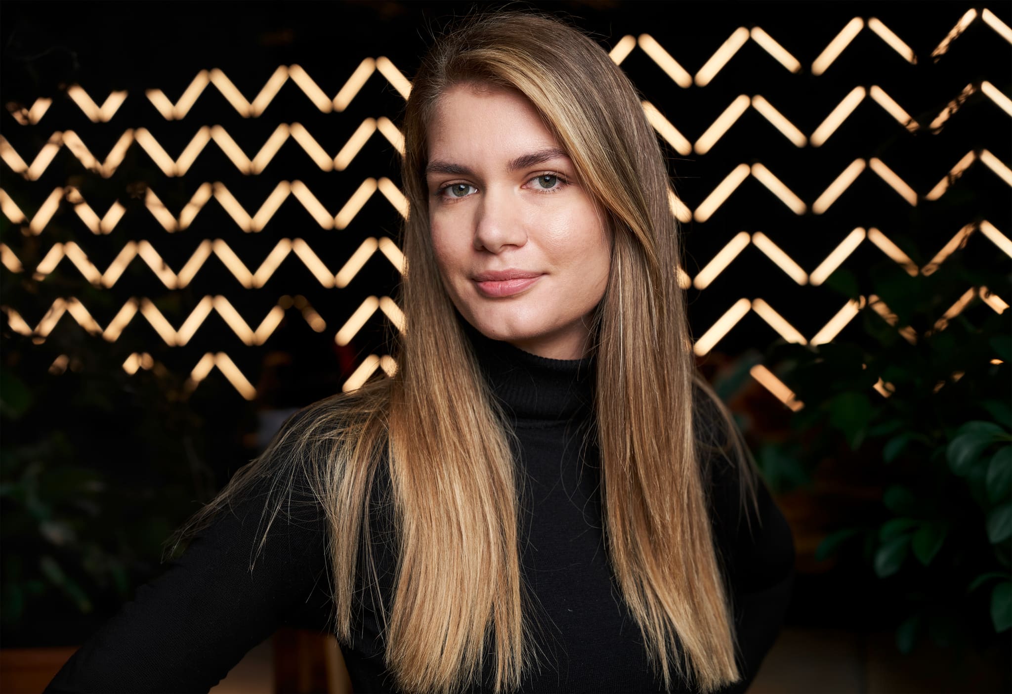 Lana Leko im schwarzen Rollkragenpullover vor einer schwarzen Wand mit Zickzack-Leuchtbändern und Pflanzen, Blick in die Kamera