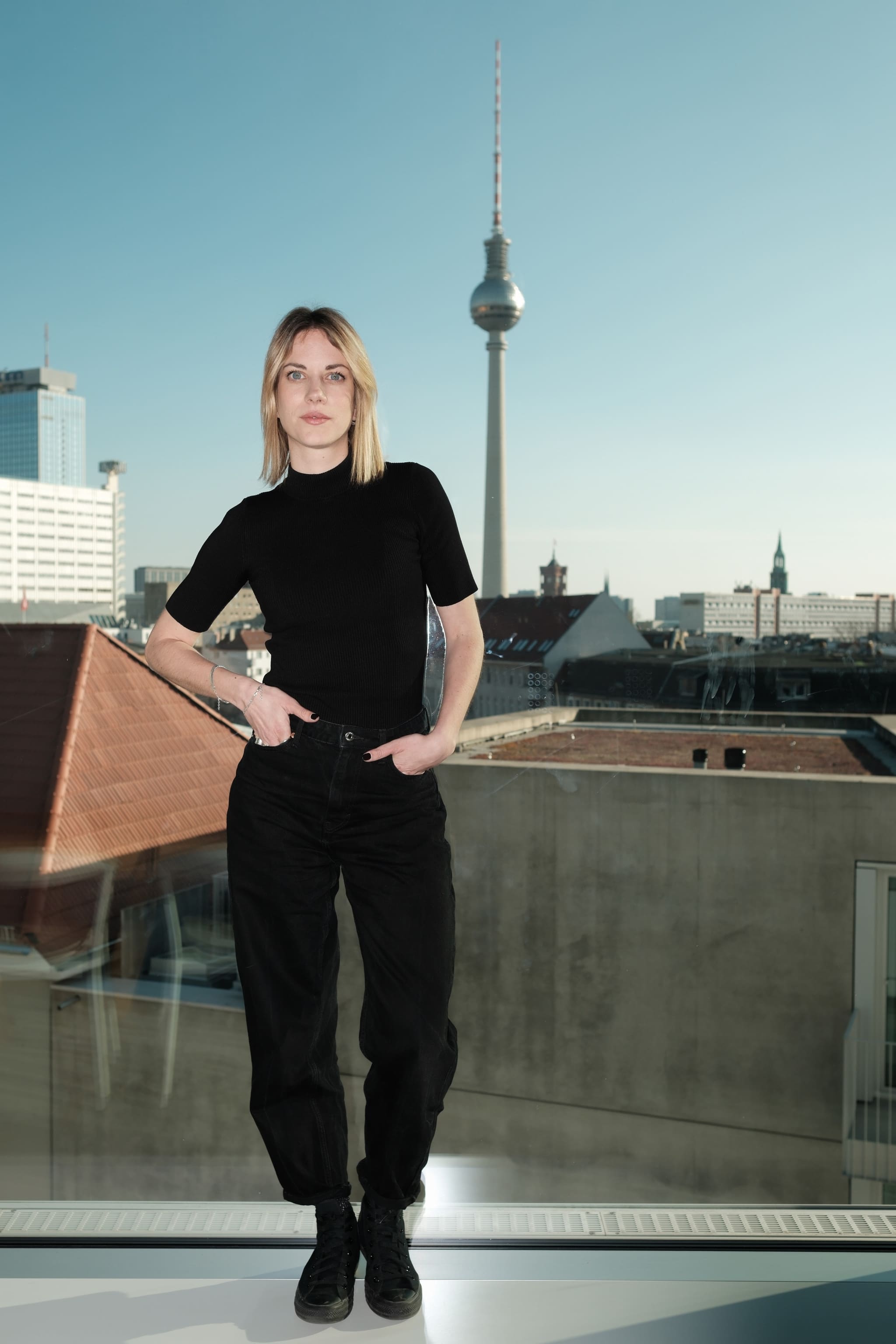 Leda Kremmyda in Schwarz vor der Fensterfront, dahinter Berliner Dächer und der Fernsehturm